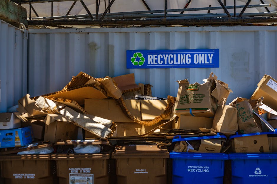 A collection of various cardboard boxes and paper packaging materials piled on top of recycling bins in an outdoor area, with some flattened and others partially crushed. The boxes are made from corru