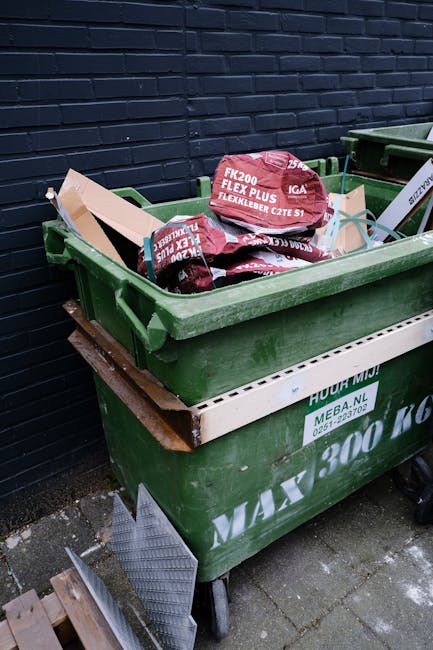 A collection of various cardboard boxes and paper packaging materials piled on top of recycling bins in an outdoor area, with some flattened and others partially crushed. The boxes are made from corru