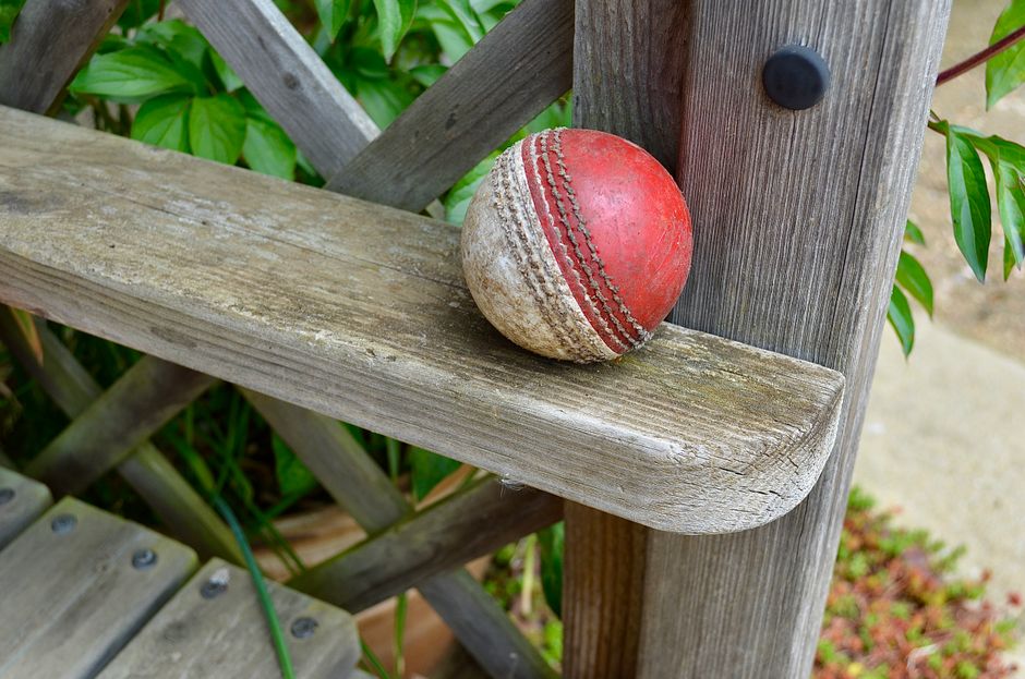 A weathered, aged wooden garden bench with visible grain and signs of wear, situated outdoors amidst greenery. Resting on the bench is a used cricket ball with a red leather surface and prominent whit